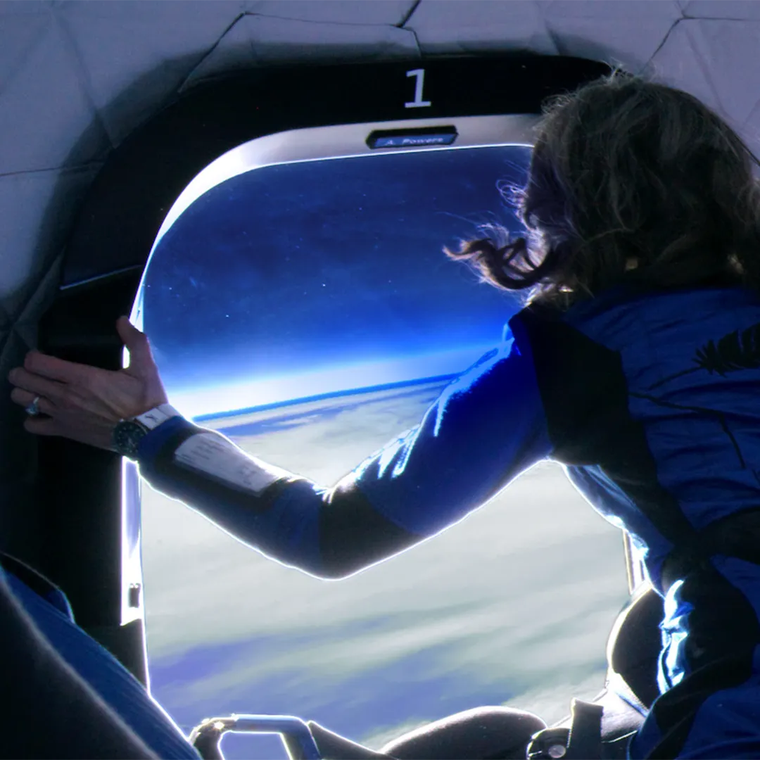 A person in a blue flight suit looks out a spacecraft window at the earth's curvature.